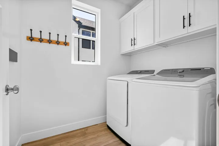 Dedicated laundry area featuring white shaker-style cabinetry with matte black hardware, a light wood-finish flooring, a single window, and a wall-mounted wooden coat rack with black hooks