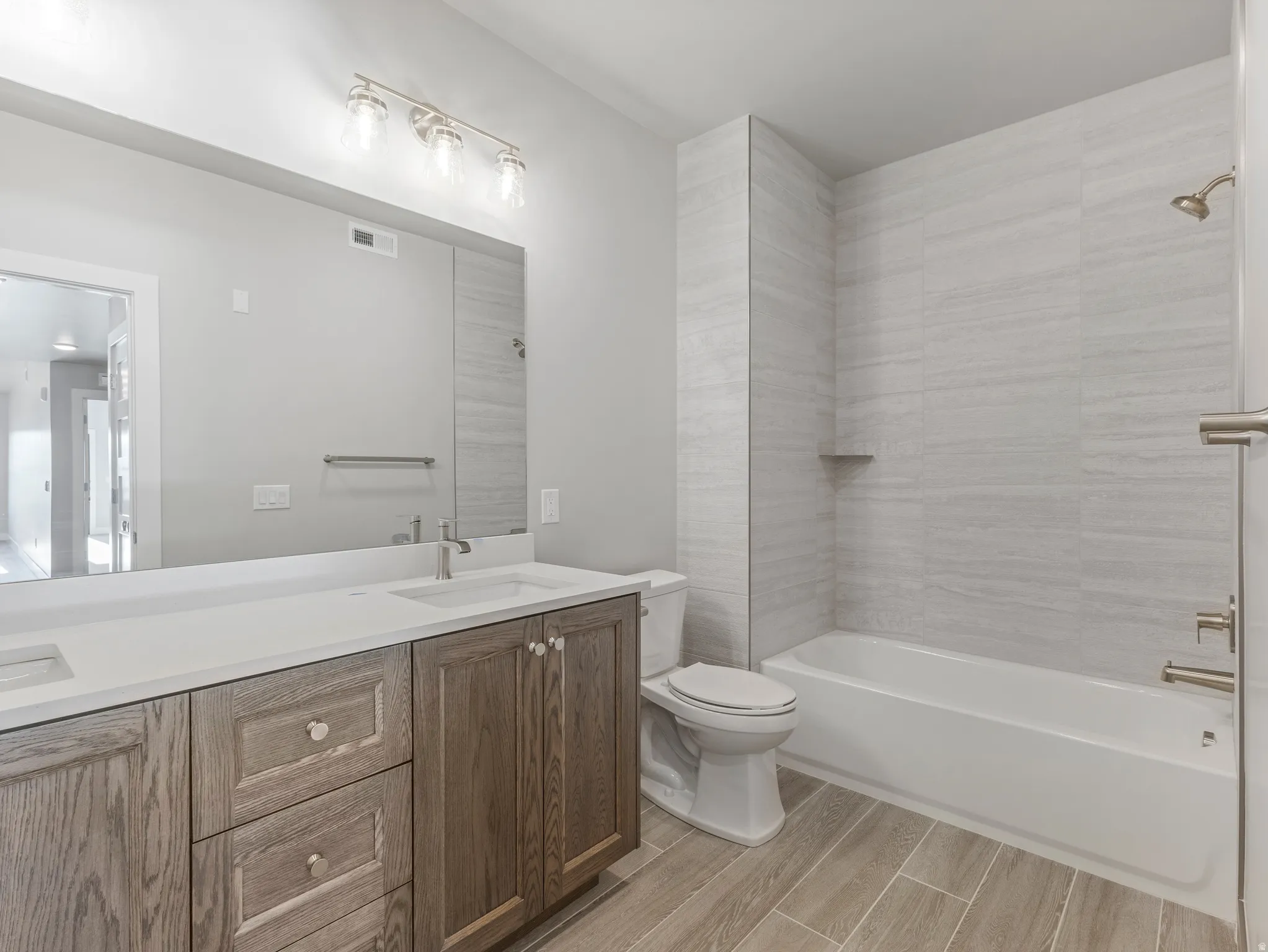 Bathroom featuring a double vanity with white countertops and wood-finish cabinetry, a large wall-mounted mirror, and a full bathtub with tiled surround