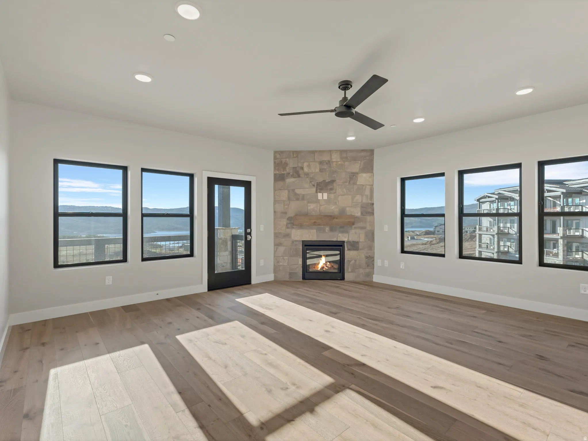 Spacious room featuring wood-finish flooring, a stone-clad fireplace, and multiple windows with black framing