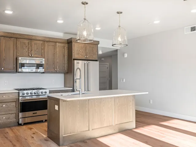 Open-concept kitchen featuring wood-finish cabinetry, stainless steel appliances, a center island with sink, and pendant lighting