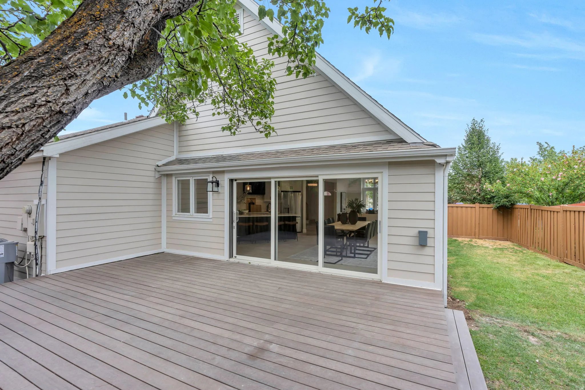 Expansive composite deck with a sliding glass door entry to the interior, featuring horizontal siding, a fenced grass area, and mature trees