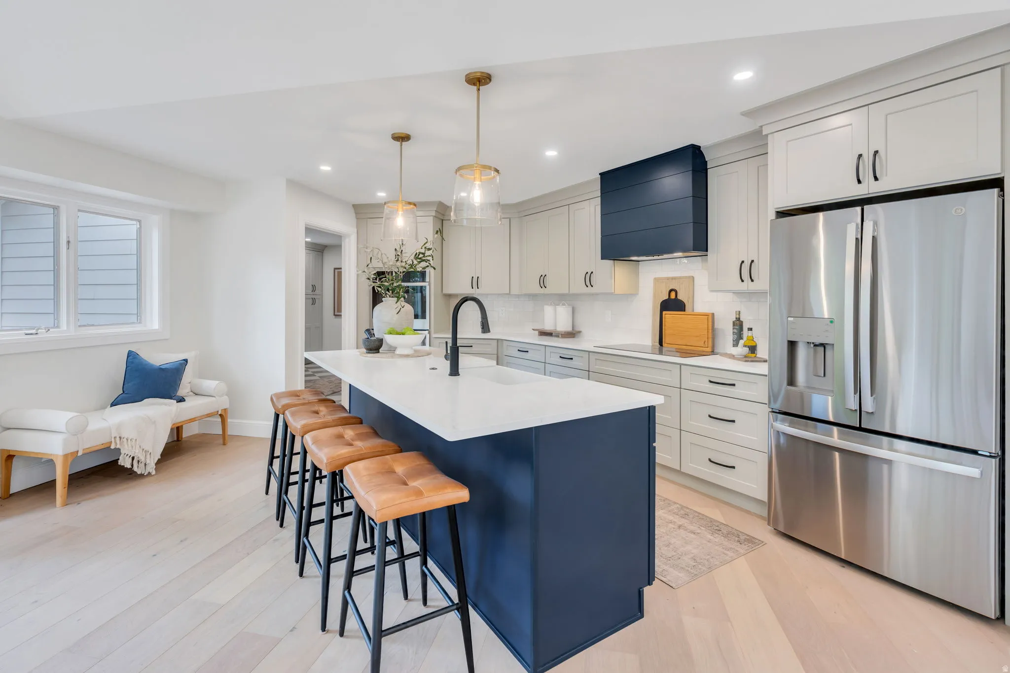 Spacious kitchen featuring a large central island with seating, stainless steel refrigerator, light wood-finish flooring, a navy range hood, and light-toned cabinetry