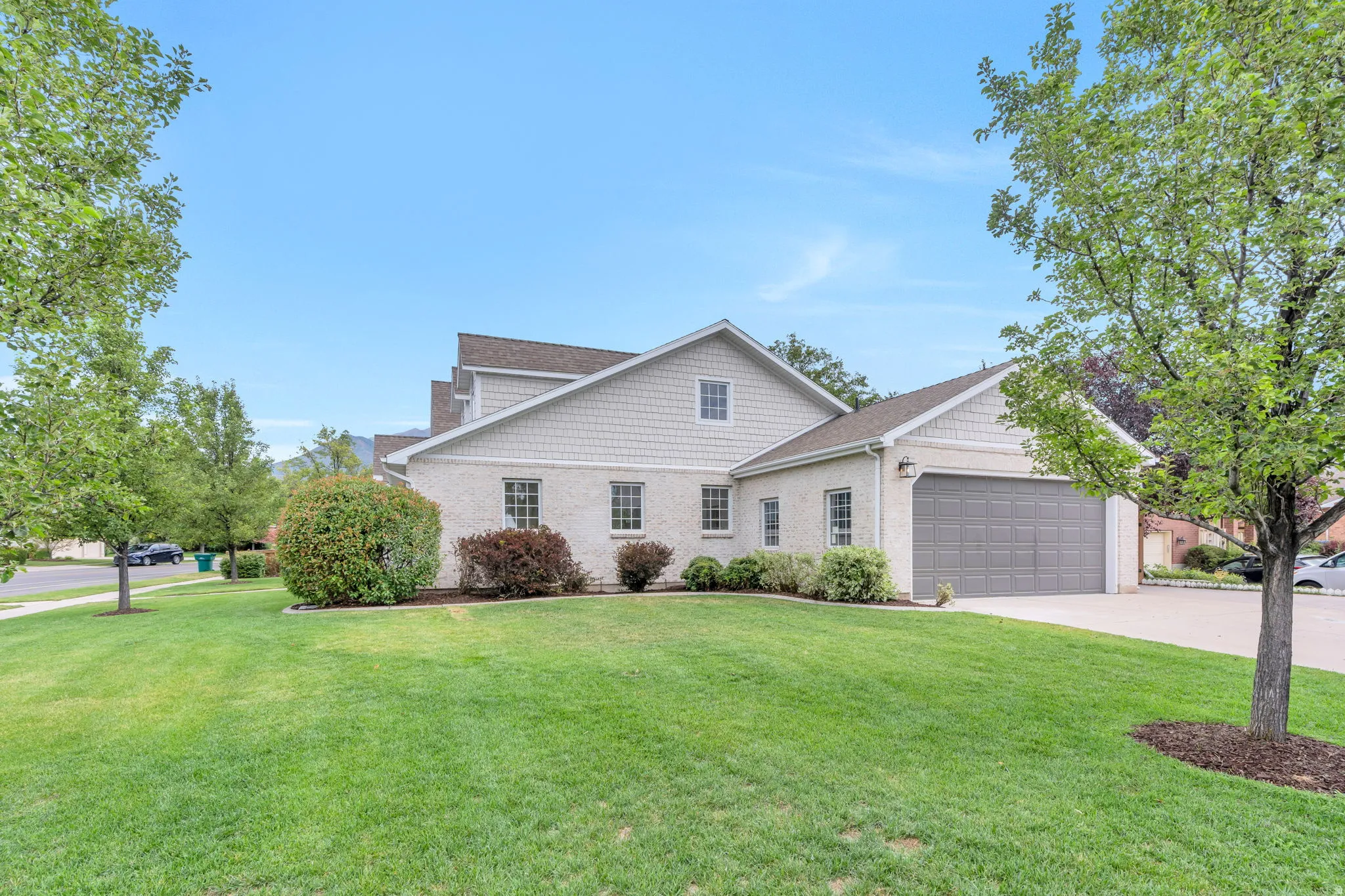 Spacious lawn with mature trees, a two-car garage, and a light-colored exterior featuring a blend of stucco and shingle siding
