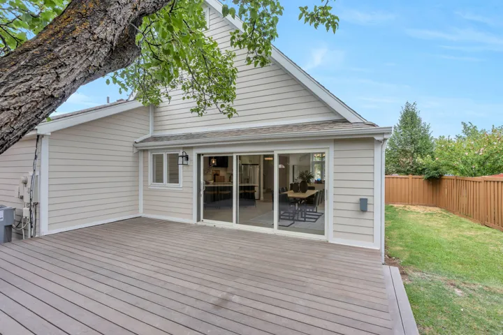 Expansive composite deck with a sliding glass door entry to the interior, featuring horizontal siding, a fenced grass area, and mature trees