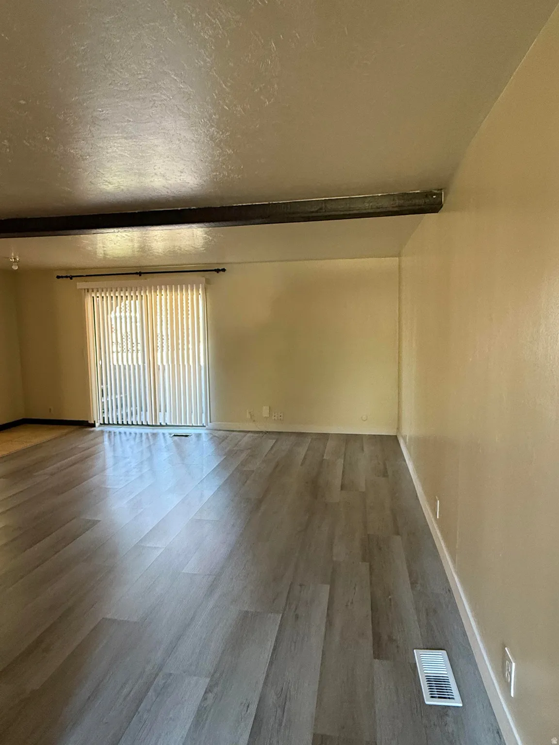 Spacious room featuring wood-finish flooring, a prominent exposed beam, and sliding glass doors with vertical blinds