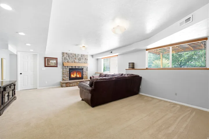 Living area featuring a stone-clad fireplace with a wood mantel, light-toned carpeting, recessed ceiling lighting, and a large window with horizontal blinds