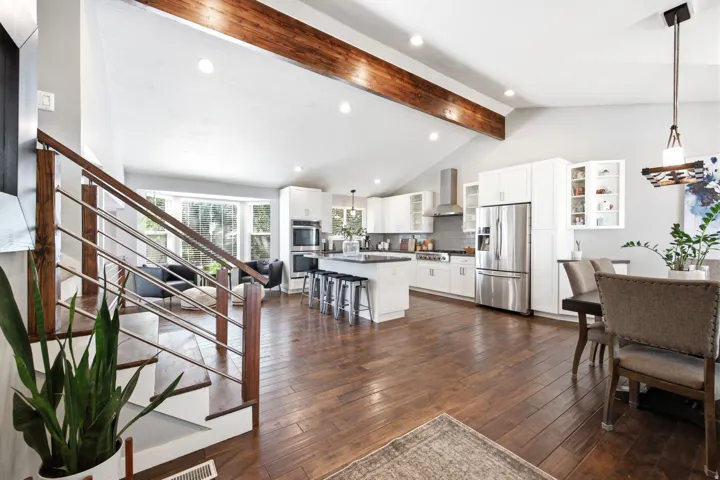 Open-concept interior featuring vaulted ceilings with exposed wood beams, wood-finish flooring, and a modern staircase with wood and metal railings
