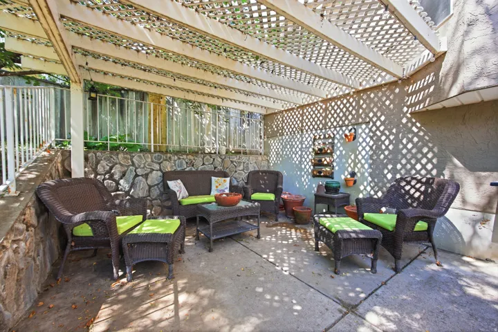 Outdoor patio area featuring a white lattice pergola, a concrete slab foundation, a stone retaining wall, stucco siding, and a white metal railing