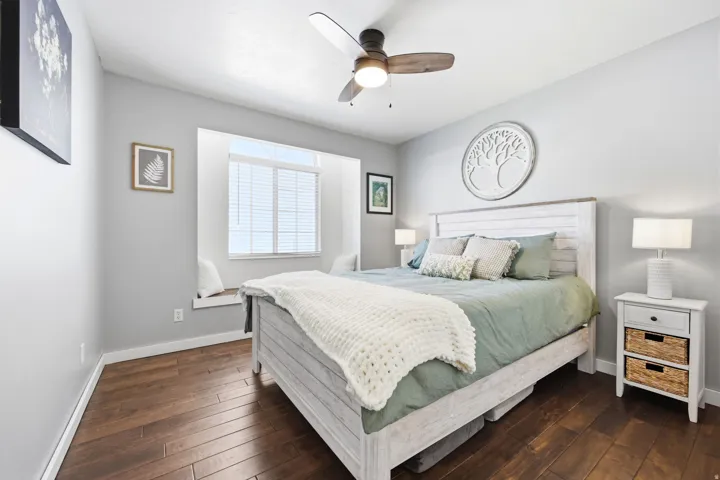 Bedroom featuring wood-finish flooring, a window seat with built-in storage, a ceiling fan with integrated lighting, a white shiplap-style headboard, and light gray wall paint