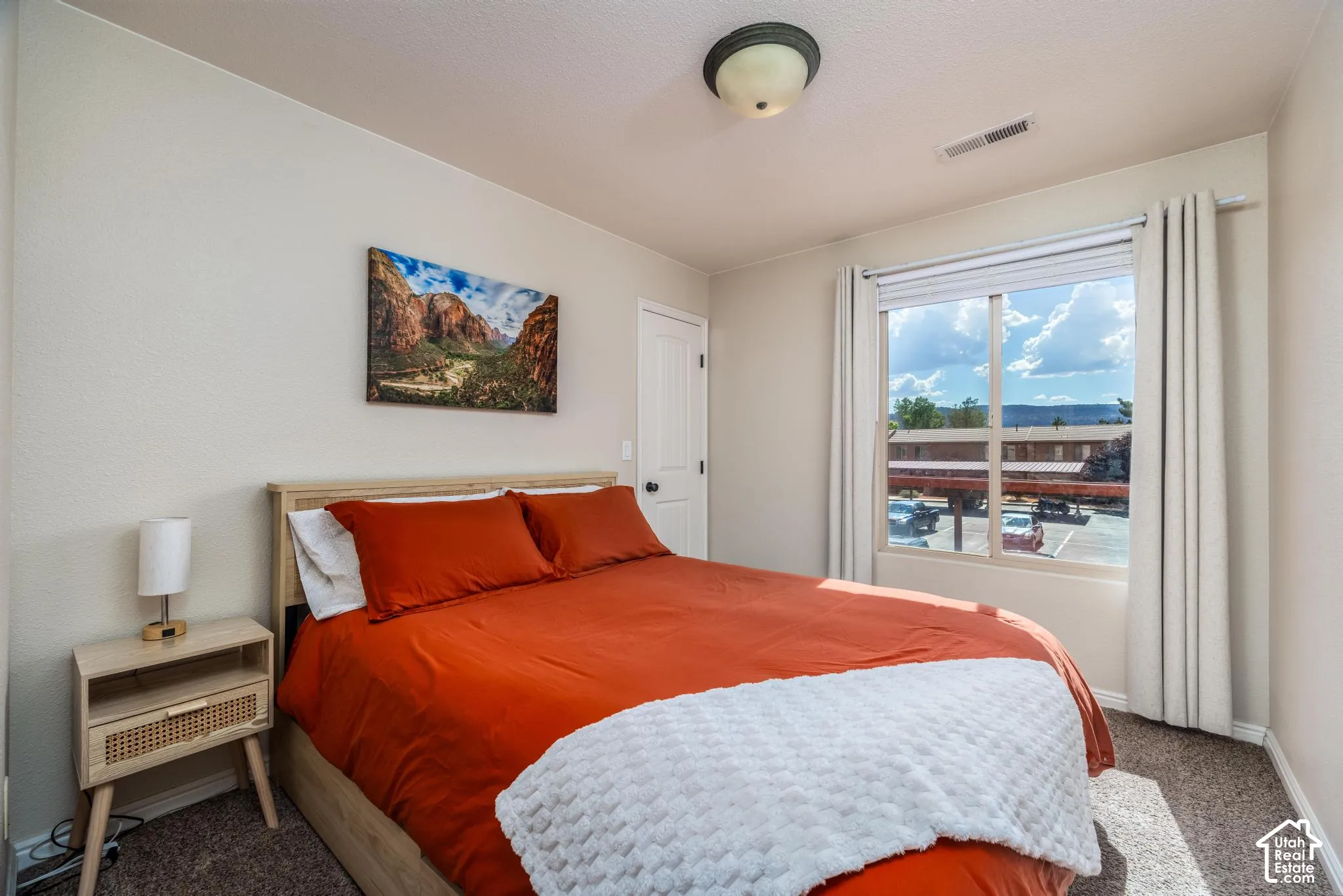 Carpeted bedroom featuring a large window with white blinds and curtains, a light-colored door, and a flush-mount ceiling light fixture