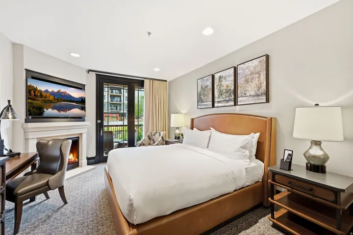 Bedroom featuring a built-in fireplace with a television above, a wood-finish desk with a leather chair, a private balcony with dark-framed double doors, a leather-upholstered bed frame, and carpeted flooring