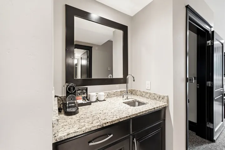 Wet bar featuring a dark wood framed mirror, stone countertop, undermount sink, and dark wood cabinetry with silver-toned hardware