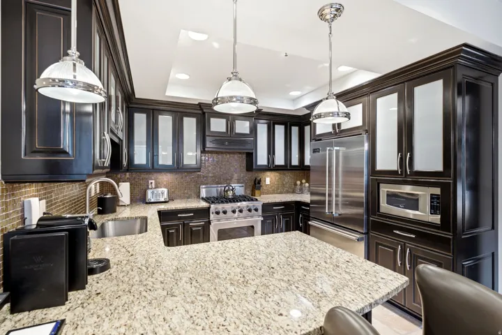 Kitchen featuring granite countertops, dark wood cabinetry with frosted glass inserts, a mosaic tile backsplash, stainless steel appliances, and pendant lighting