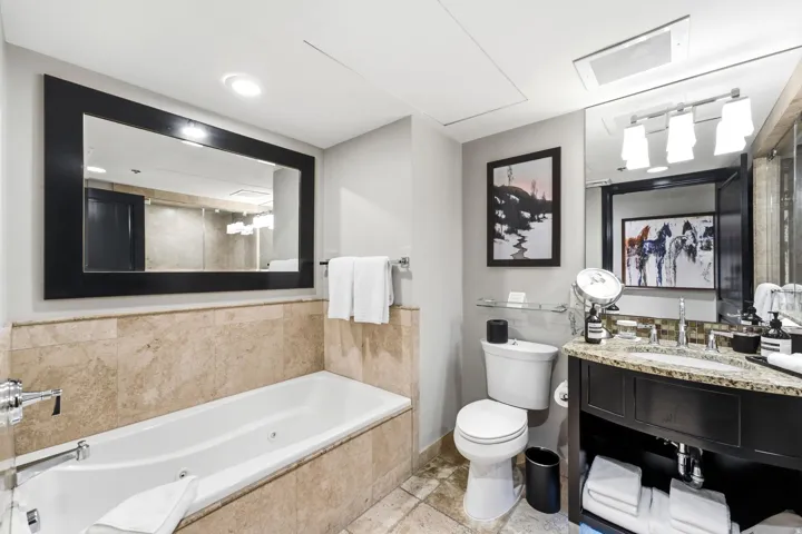 Bathroom featuring a built-in jetted tub surrounded by natural stone tiling, a dark wood-finish vanity with an undermount sink and stone countertop, a wall-mounted mirror with integrated lighting, and a white toilet