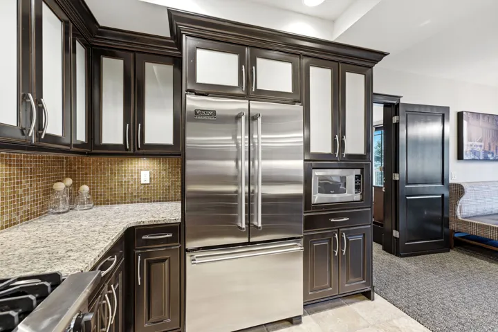 Gourmet kitchen featuring a stainless steel French door refrigerator, built-in microwave, dark wood cabinetry with frosted glass inserts, speckled countertops, and a mosaic tile backsplash