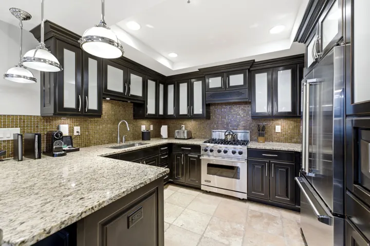 Kitchen featuring dark cabinetry with glass-front uppers, light-toned stone countertops, a mosaic tile backsplash, and stainless steel appliances