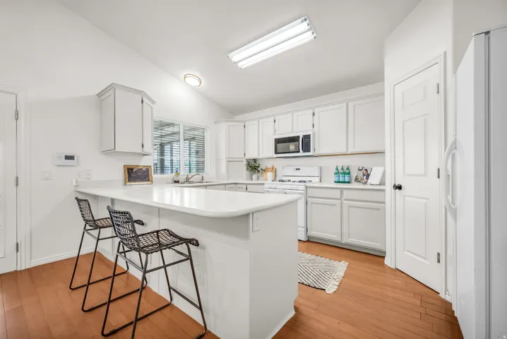 Kitchen featuring an eat-at peninsula, white cabinetry, light countertops, wood-finish flooring, and white appliances