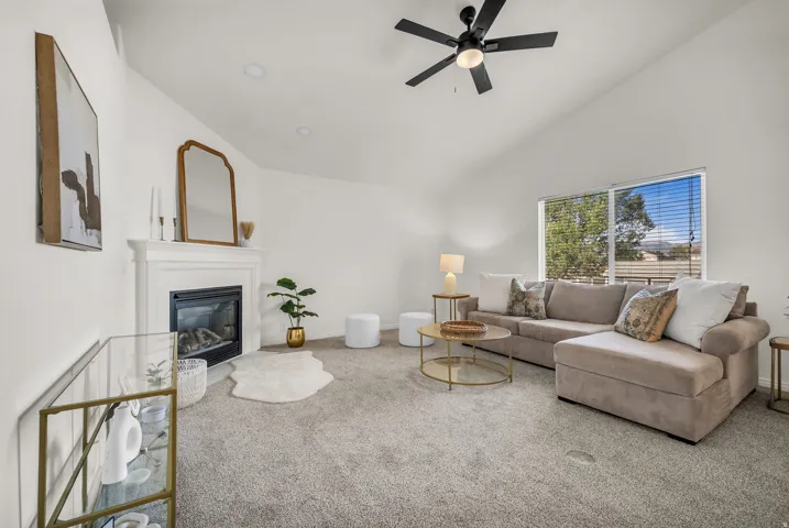 Spacious living area featuring vaulted ceilings, a modern fireplace with white mantel, broad window with horizontal blinds, and a black ceiling fan with integrated lighting