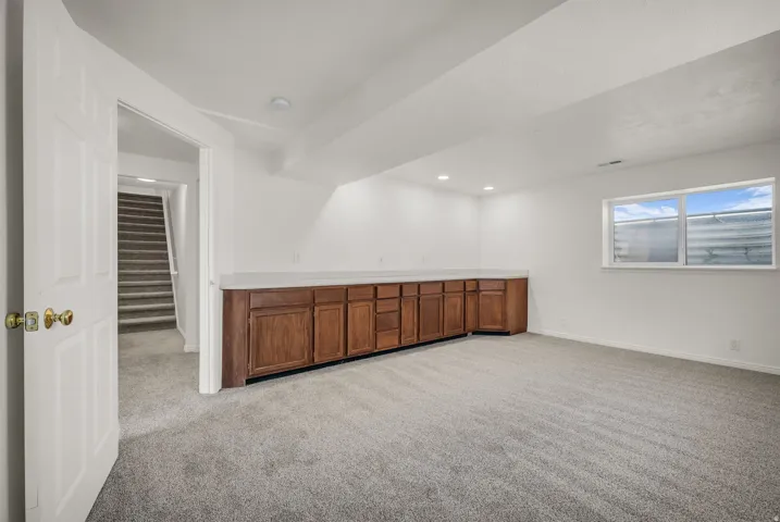 Spacious room featuring light gray carpet flooring, white walls, recessed lighting, and a long built-in wood-finish cabinet with a light-toned countertop