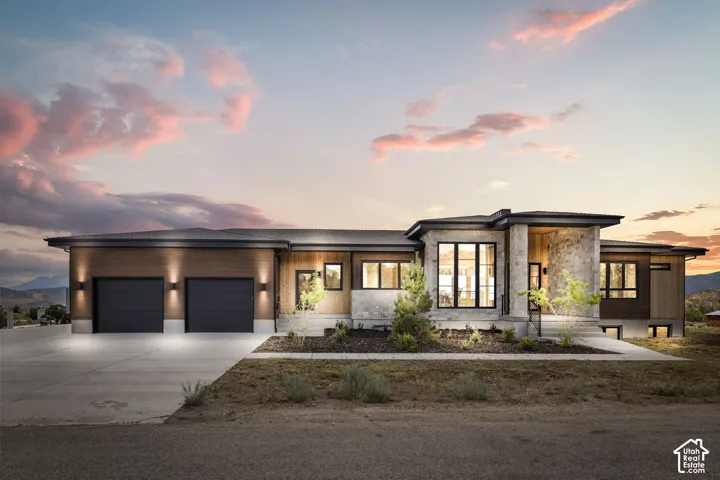 Contemporary exterior featuring stone and wood-look siding, a prominent two-car garage, large black-framed windows, and integrated exterior lighting