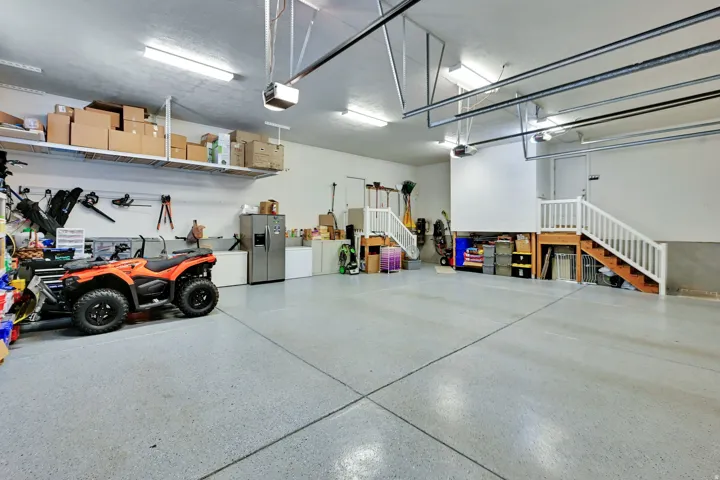 Spacious garage featuring a light-colored epoxy floor, ample overhead lighting, built-in shelving, two sets of interior stairs, and a partial second level