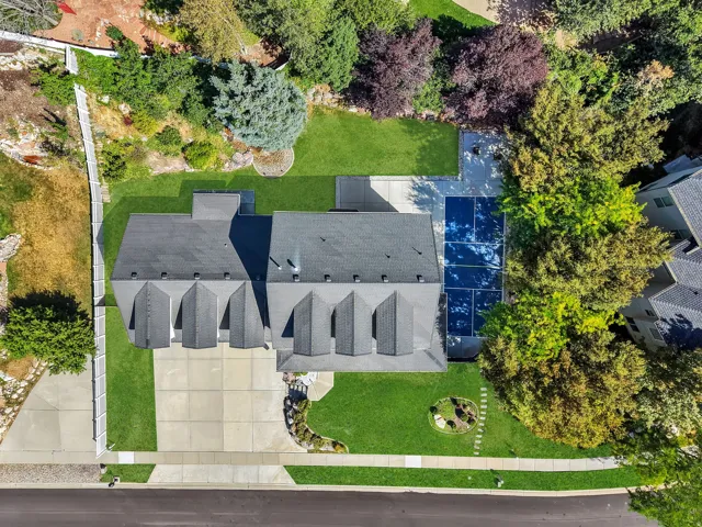 Aerial view of a residential property featuring a large asphalt shingle roof with multiple dormers, extensive concrete driveway, mature landscaping with manicured lawns, a fenced backyard, and solar panels