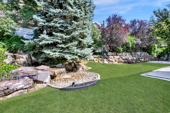 Expansive backyard featuring a multi-tiered rock waterfall, a manicured lawn, mature evergreen tree, and a stone retaining wall