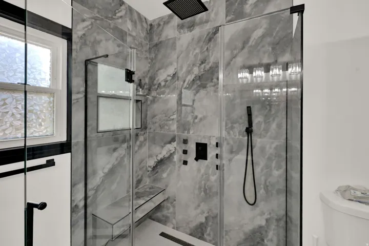 Walk-in shower with gray marbled wall tiling, black matte fixtures, a built-in bench, a rain showerhead, and a handheld shower wand