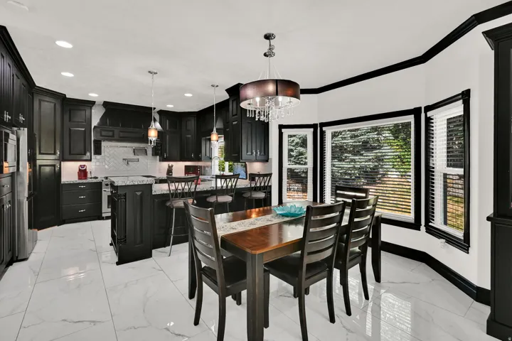 Modern kitchen and dining area featuring black cabinetry, white marble-look tile flooring, bay windows, a central island with bar seating, and a decorative chandelier