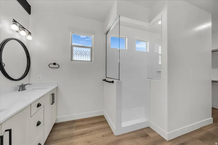 Bathroom featuring a glass-enclosed shower with white subway tile surround, a single vanity with a white countertop and light wood-finish cabinetry, a black framed oval mirror, and wood-finish flooring
