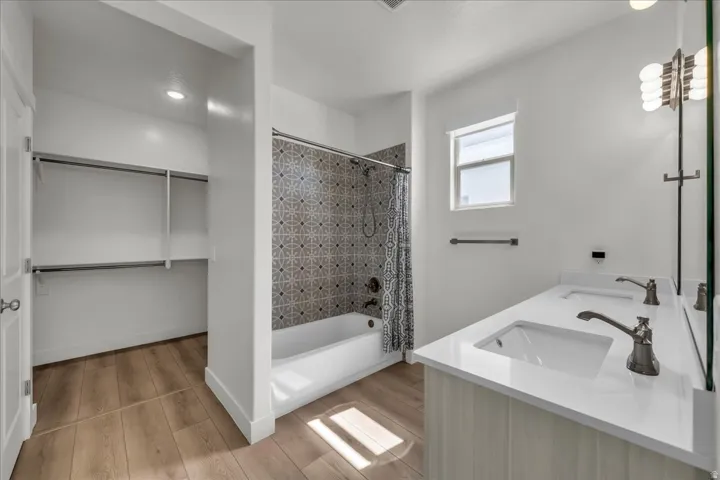 Bathroom featuring a double vanity with white countertops, a tub/shower combination with patterned tile surround, wood-finish flooring, and a walk-in closet with built-in shelving