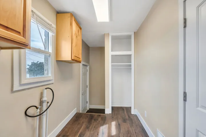 Laundry area featuring wood-finish flooring, two upper wood cabinets, and a built-in shelving unit with a hanging rod