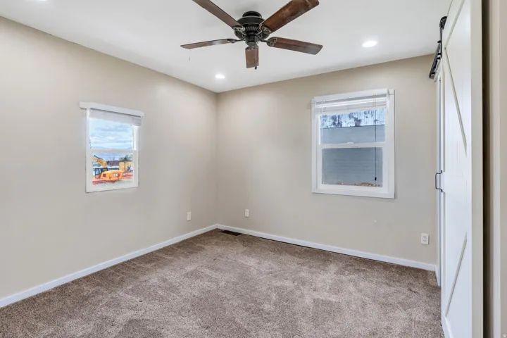 Carpeted interior room featuring recessed lighting, a ceiling fan with wood-finish blades, two windows with blinds, and a white barn-style sliding door