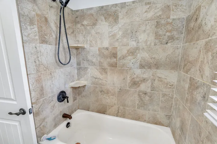 Bathtub with stone-finish tile surround, integrated shower shelves, dark bronze shower fixtures, and white paneled door