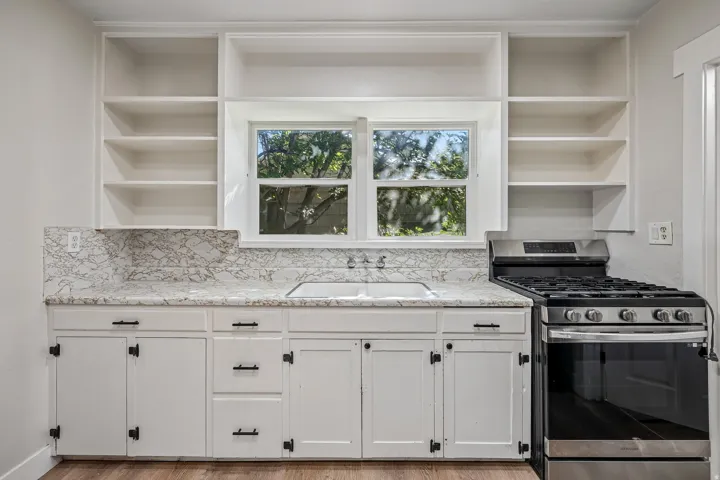 White shaker-style cabinetry with matte black hardware, integrated window over the sink, built-in open shelving, and a stainless steel range