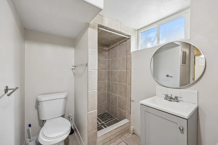 Tiled shower stall with a mosaic-patterned floor, white toilet, vanity with a white countertop, and a circular mirror