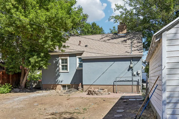 Rear exterior featuring gray stucco siding, a shingle roof, a brick chimney, and a stone-paver fire pit