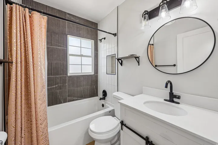 Modern bathroom featuring a ceramic vessel sink with matte black faucet and fixtures, white quartz countertop, circular framed mirror with chrome hardware, dual vanity lighting, and an enclosed shower-tub combination with textured tile surround and coordi