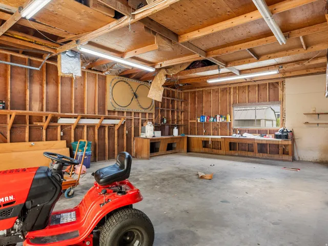 Spacious garage interior featuring exposed wood framing, concrete flooring, fluorescent lighting, built-in shelving, and a wood-finish workbench with storage