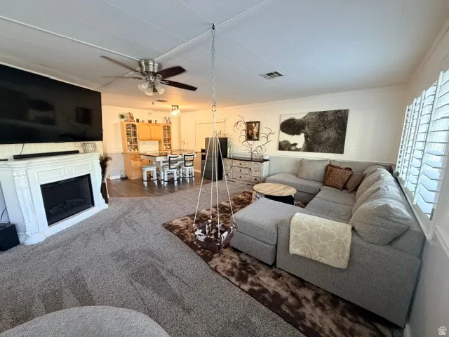 Spacious living area featuring a decorative fireplace with a mantel, carpet flooring, plantation shutters, and a ceiling fan