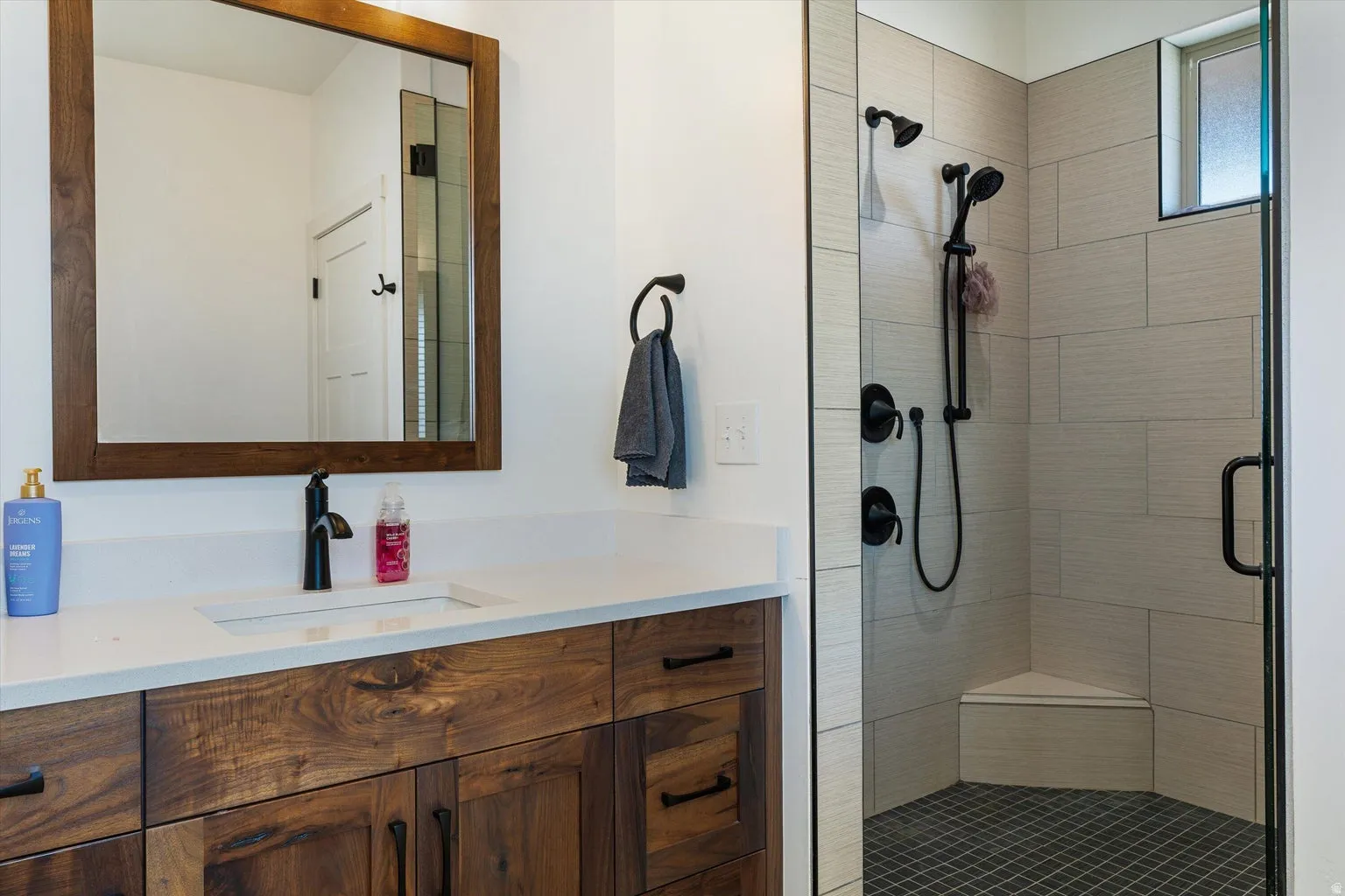 Walk-in shower with light-toned rectangular tile surround, black grid-patterned floor tile, and a built-in corner bench
