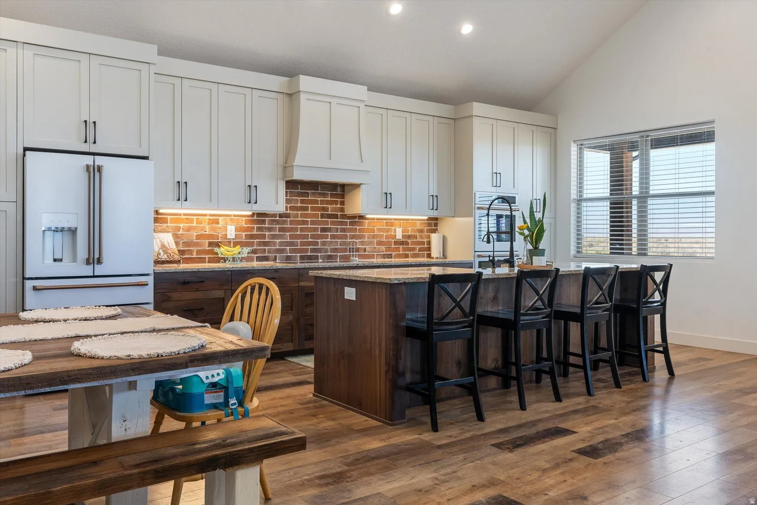 Spacious kitchen featuring a vaulted ceiling, wood-finish flooring, white upper cabinetry, a brick-patterned backsplash, and a large central island with seating