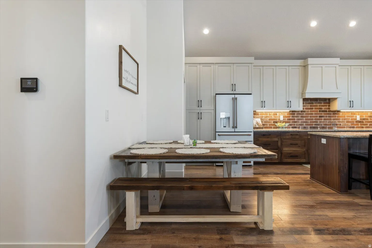 Open-concept kitchen with wood-finish flooring, white upper cabinetry, a brick-style backsplash, stainless steel refrigerator, and a kitchen island with seating