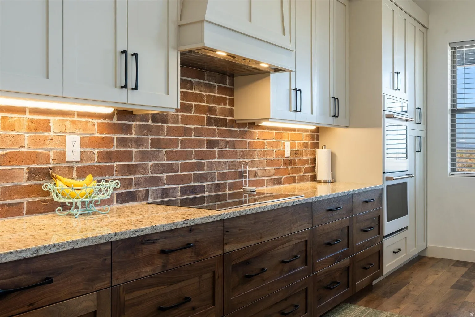 Kitchen featuring a brick backsplash, light upper cabinetry, dark wood-finish lower cabinetry, granite countertops, and built-in appliances