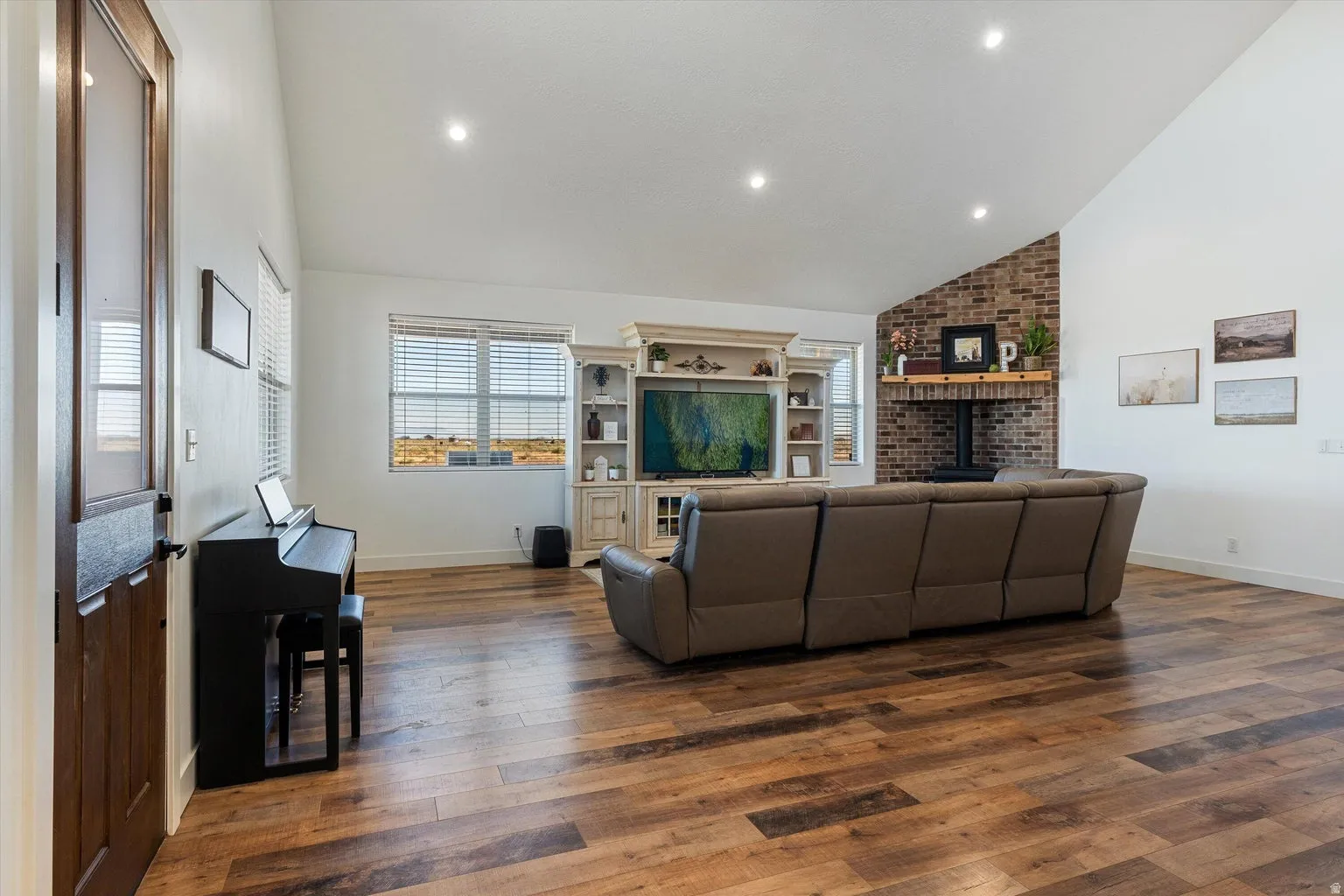 Spacious room featuring vaulted ceilings, recessed lighting, wood-finish flooring, a brick-faced fireplace with a wood mantel, and multiple windows with horizontal blinds