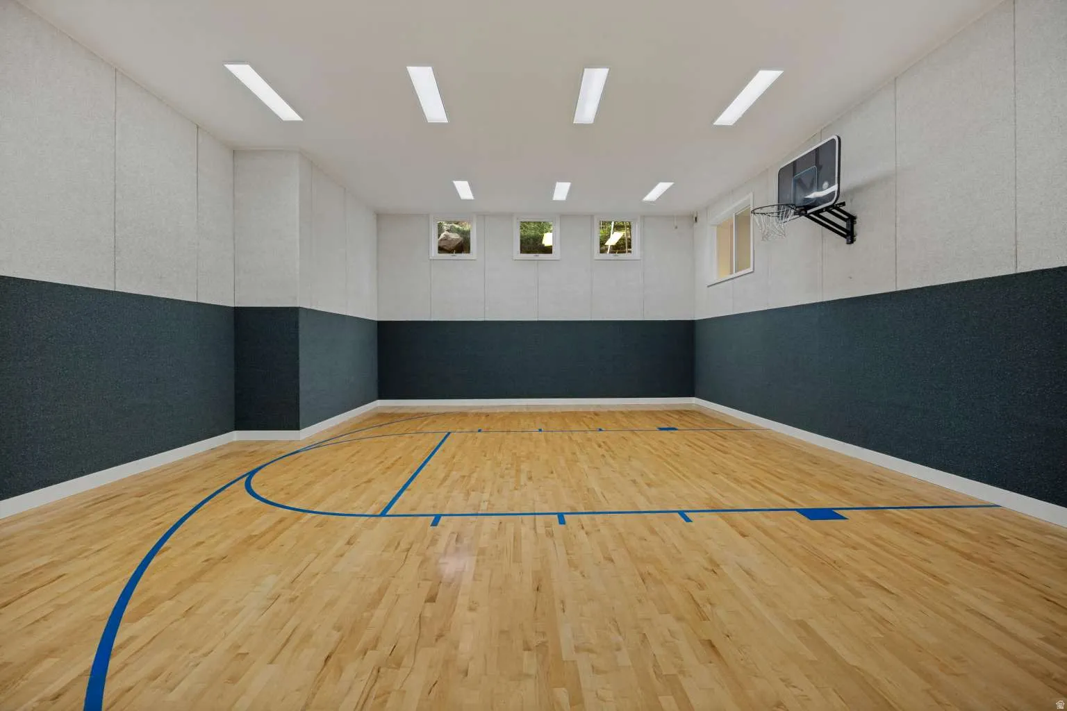 Indoor sports court featuring hardwood flooring with court markings, a mounted basketball hoop, three windows, and overhead strip lighting