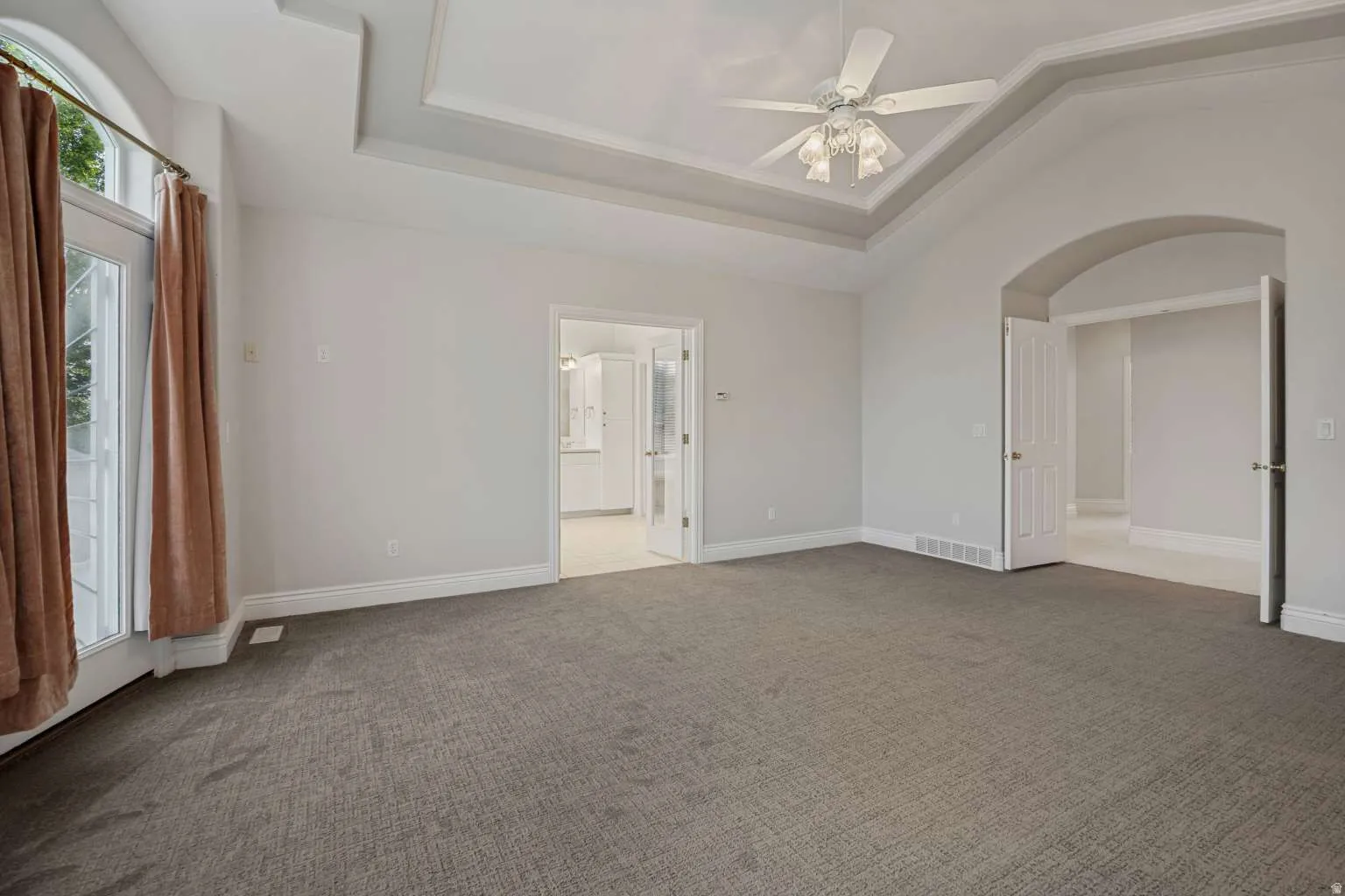 Tray ceiling with a ceiling fan, arched doorways, and light-colored carpeting