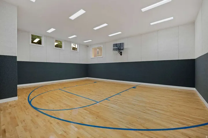 Indoor sports court featuring wood-finish flooring with blue court lines, a mounted basketball hoop, and multiple recessed rectangular light fixtures