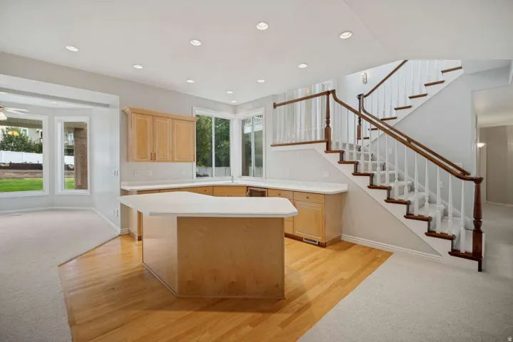 Open-concept living area featuring wood-finish flooring, a kitchen island with light-toned cabinetry, a bay window, recessed lighting, and a staircase with wood railings