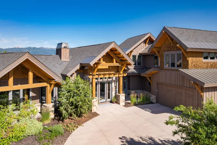 Architectural residence featuring a multi-gabled roofline, mixed material siding, exposed timber framing, stone accents, and a paved driveway with integrated landscaping